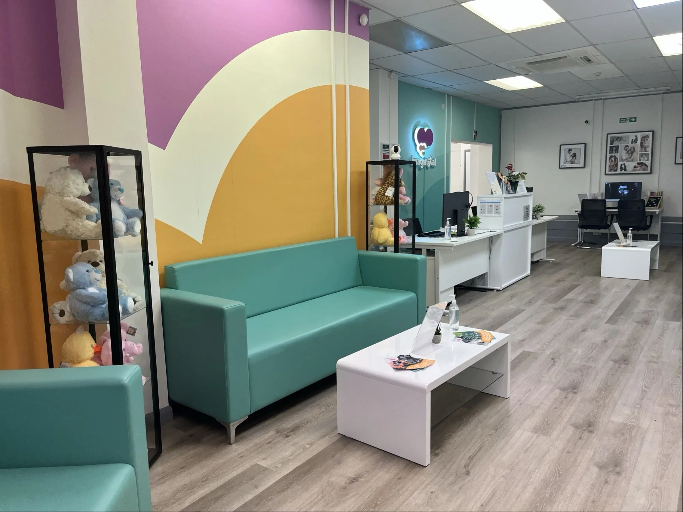 Imaginatal clinic's interior showing a bright reception area with teal sofas, baby heartbeat bears on display, modern decor, and the ultrasound viewing area in the background.