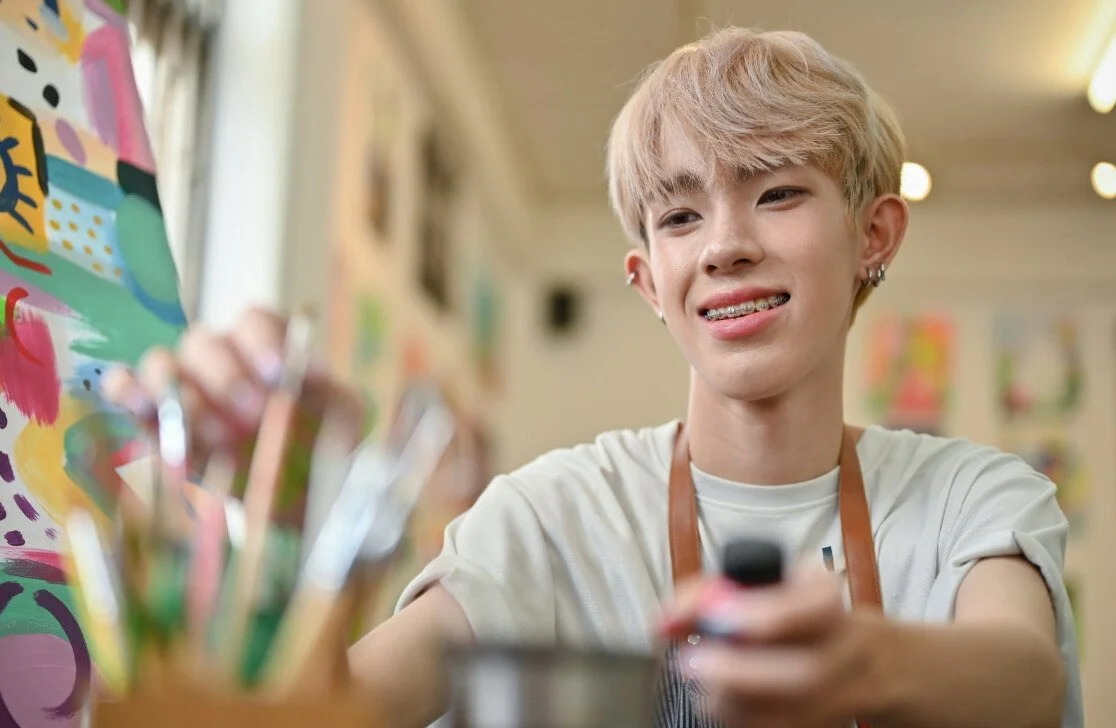 Young person with short blonde hair and braces, wearing a white shirt and apron, smiling in an art studio with colorful artwork on the walls.