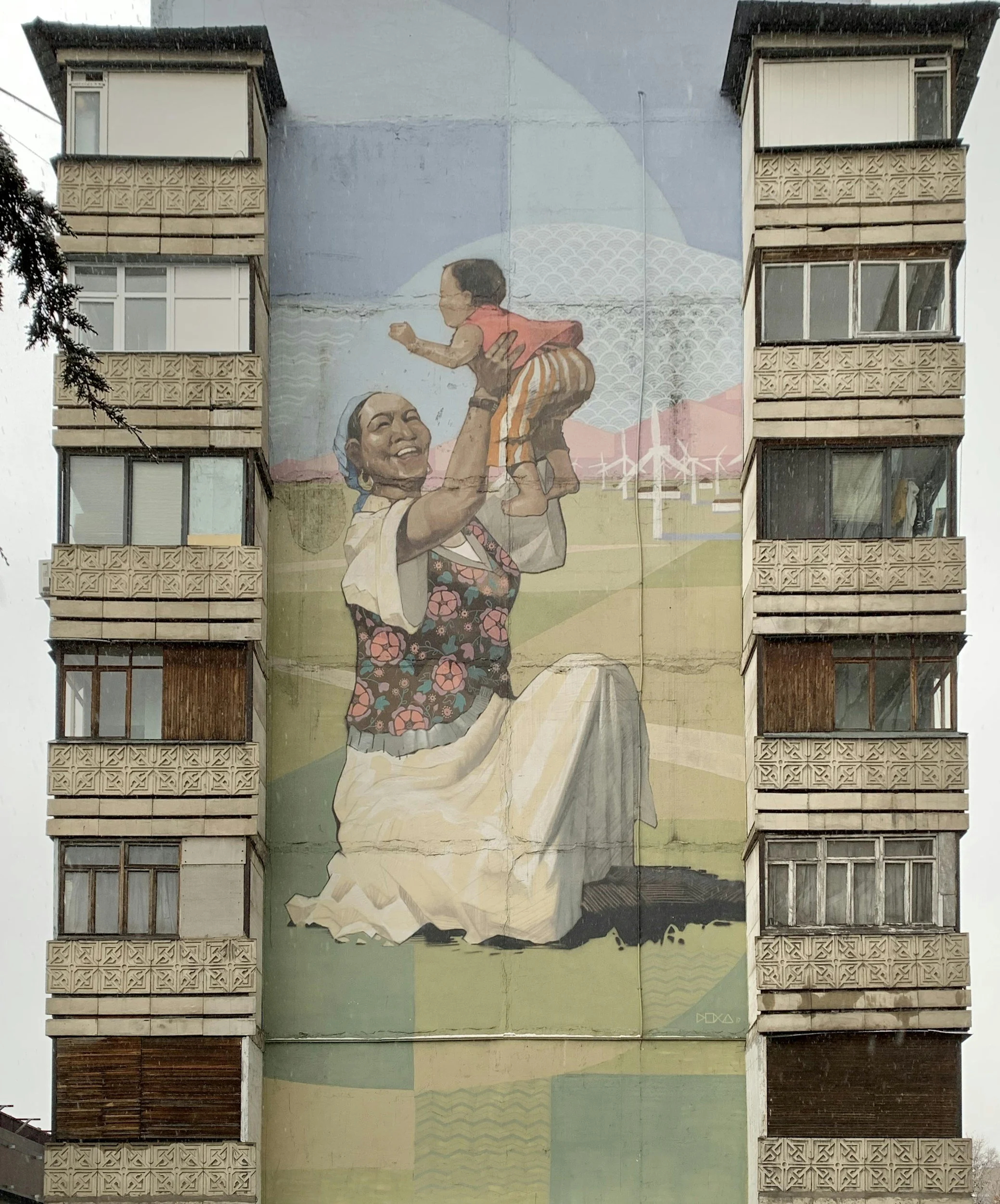 A mural on the side of a multi-story building depicting a smiling woman holding up a child against a background of wind turbines and mountains.