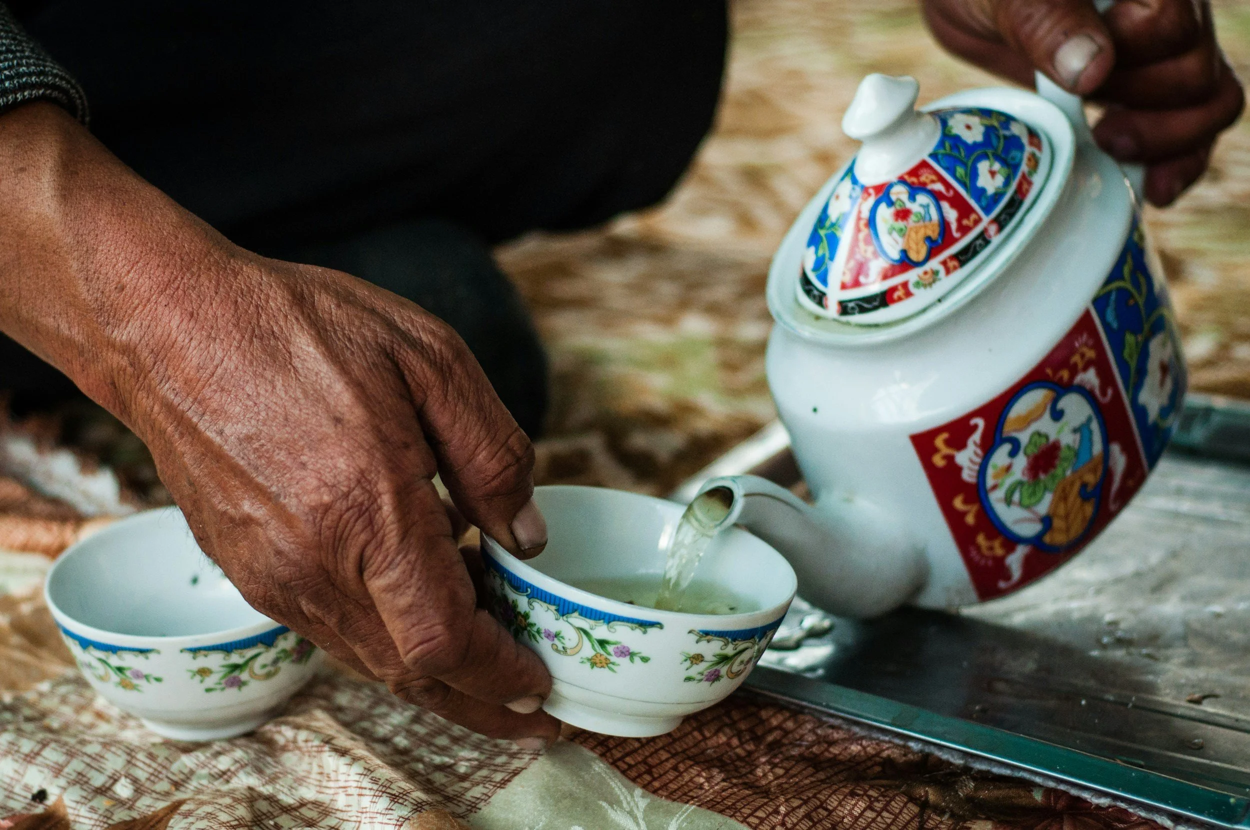 Elderly person pouring tea from a decorated teapot into a small floral teacup on a textured cloth.