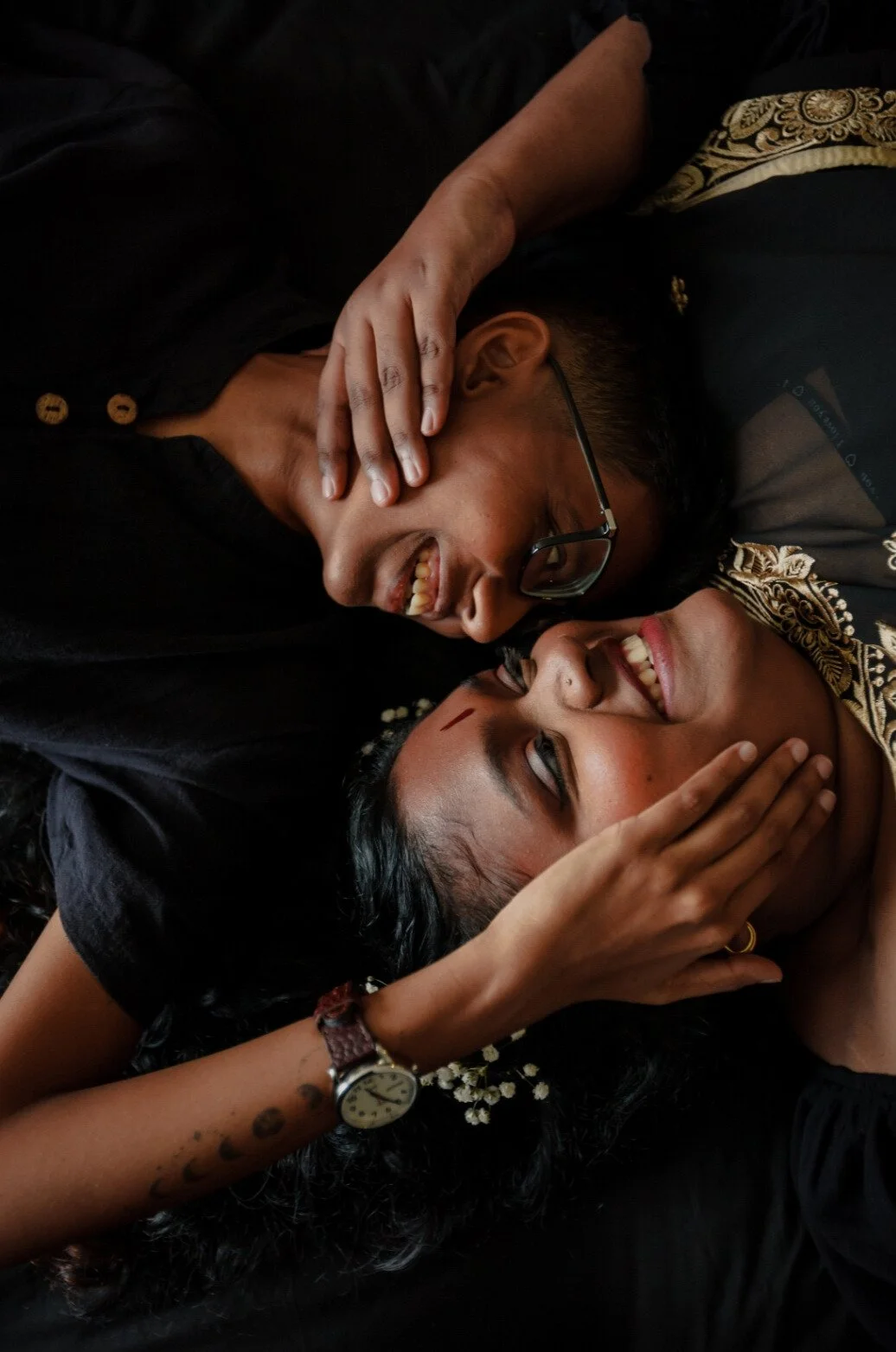 Two people are lying down, smiling, touching faces, and holding hands, wearing dark clothing and jewelry.