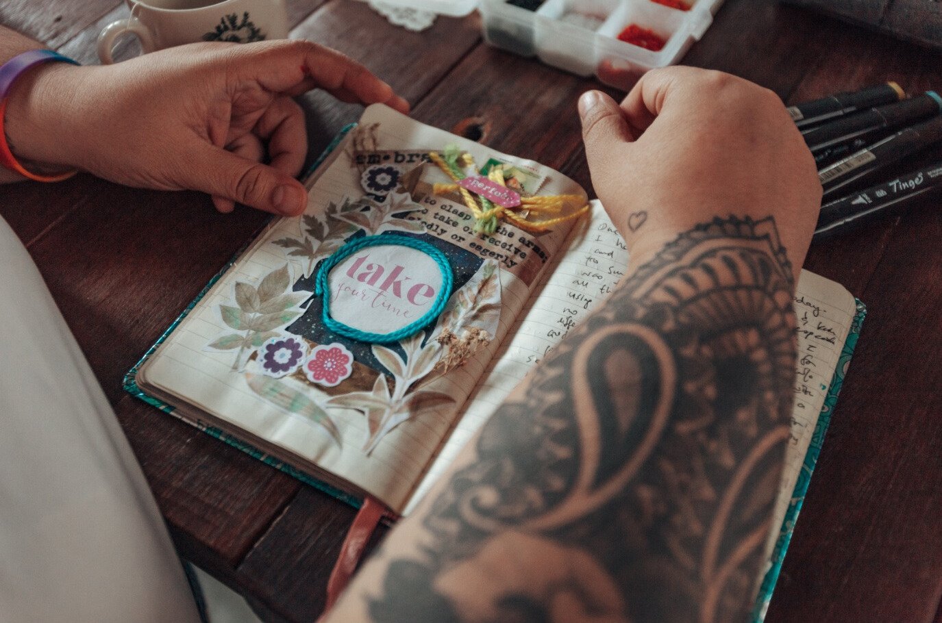 A person with tattoos on their arm is decorating a journal page with colorful stickers, including one that says 'Take your time' inside a blue rope circle. The person is writing in the journal, which is open on a dark wooden table, with some pens and small containers of art supplies nearby.