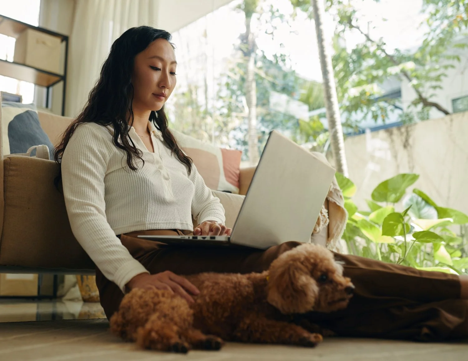 Woman with dark long hair sitting on the floor working at a laptop, petting a puppy with her right hand.
