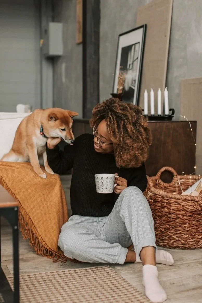 Woman with curly hair sitting on the floor, relaxed, holding a cup and petting a dog next to her.