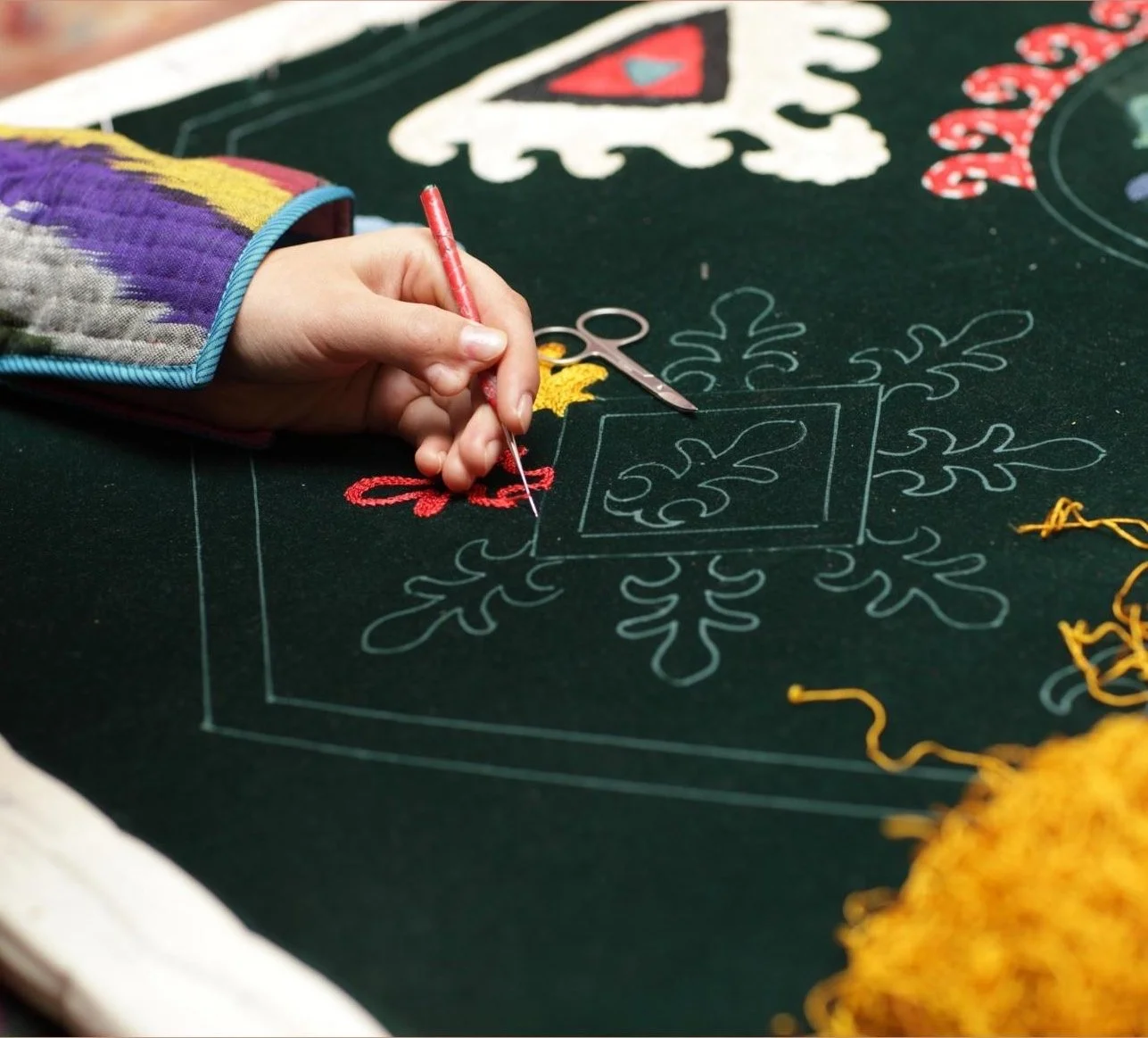 A person creating an intricate  design on green fabric using colored embroidery thread and a needle, with some embroidery scissors nearby.