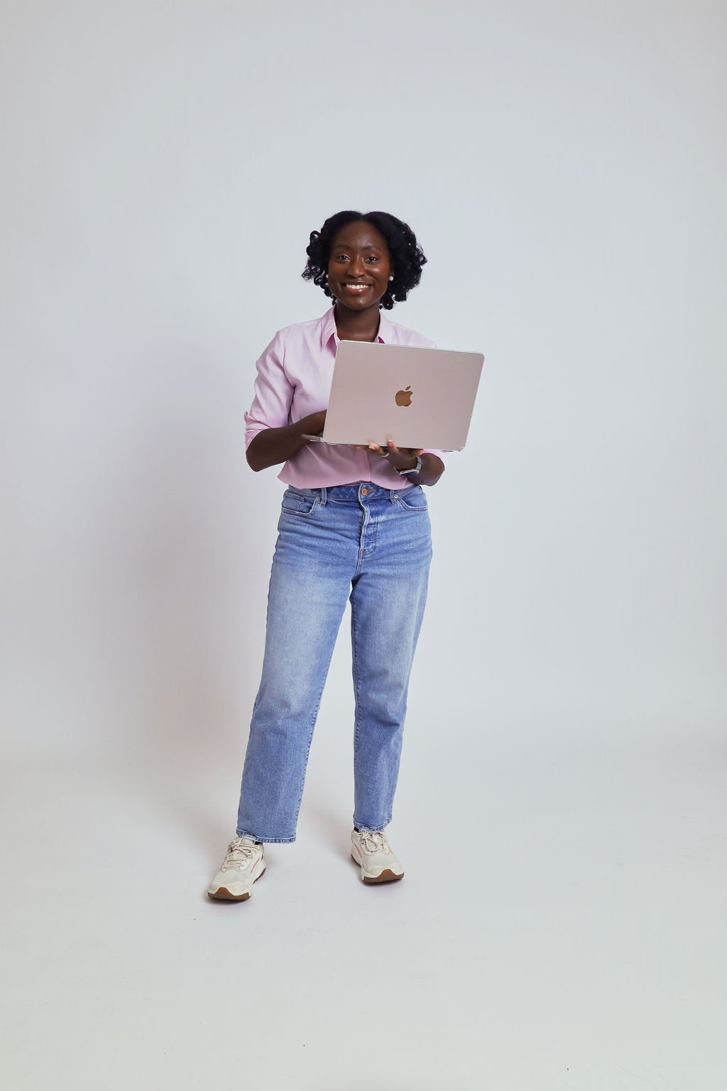 A young woman with curly dark hair smiling and holding a silver laptop while standing against a plain white background.