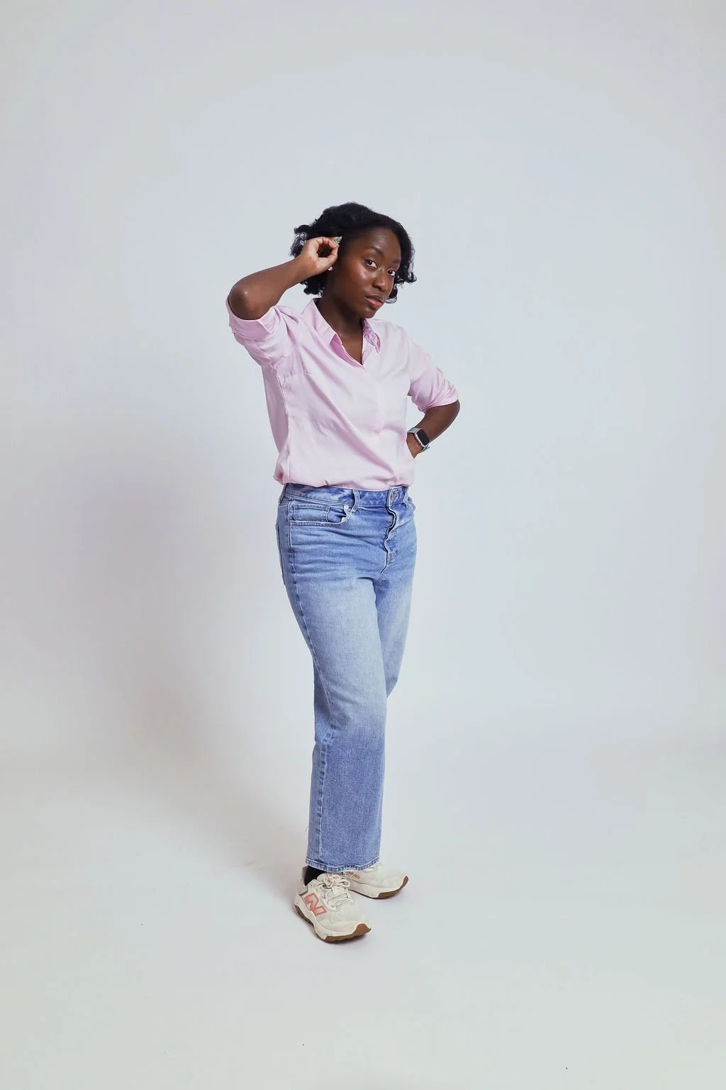 Woman with dark curly hair wearing pink shirt, blue jeans, sneakers, standing against plain light background.