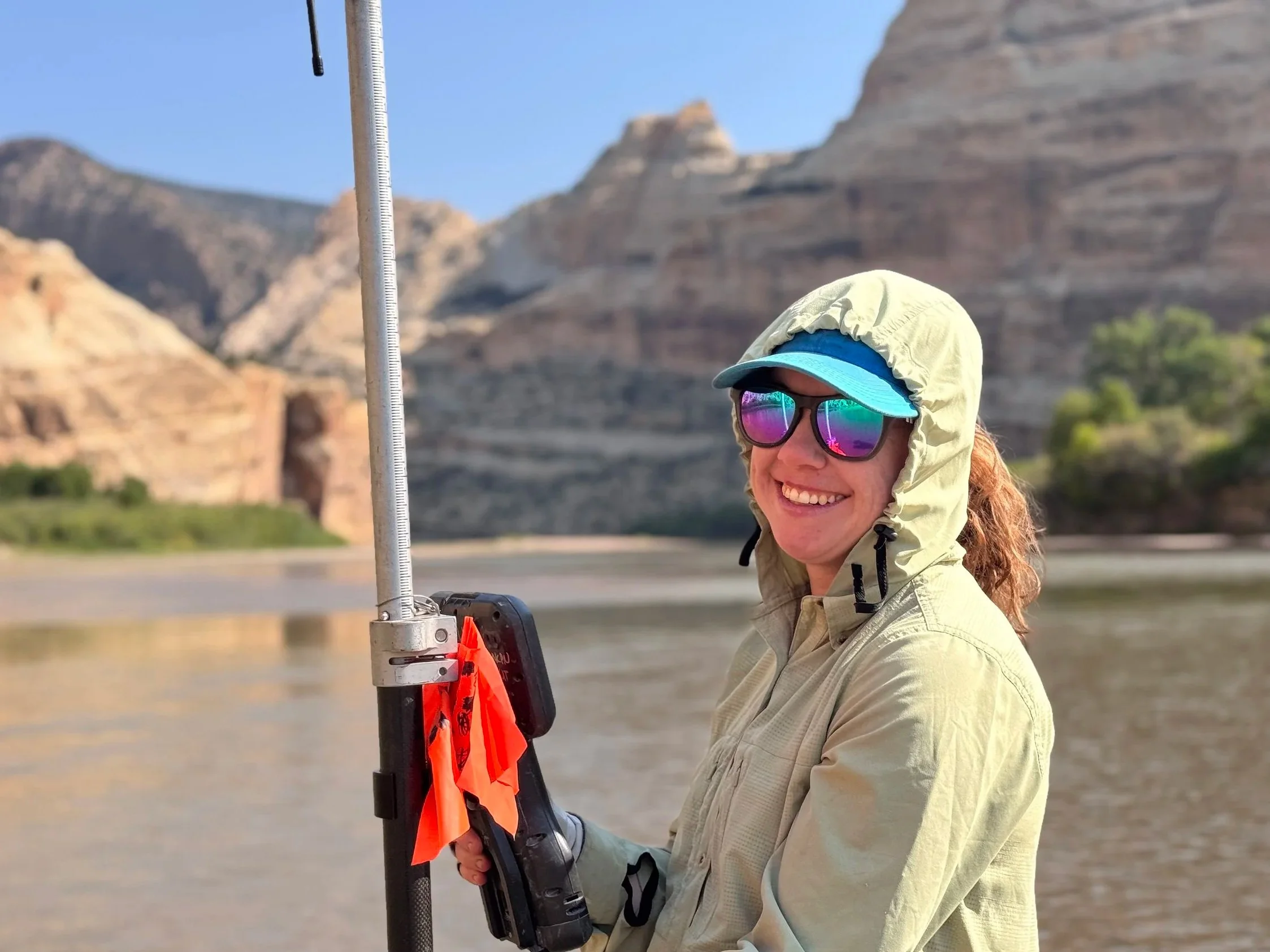 A photo of a woman wearing a sun hoodie and sunglasses holding surveying equipment by a river with rock formations in the background
