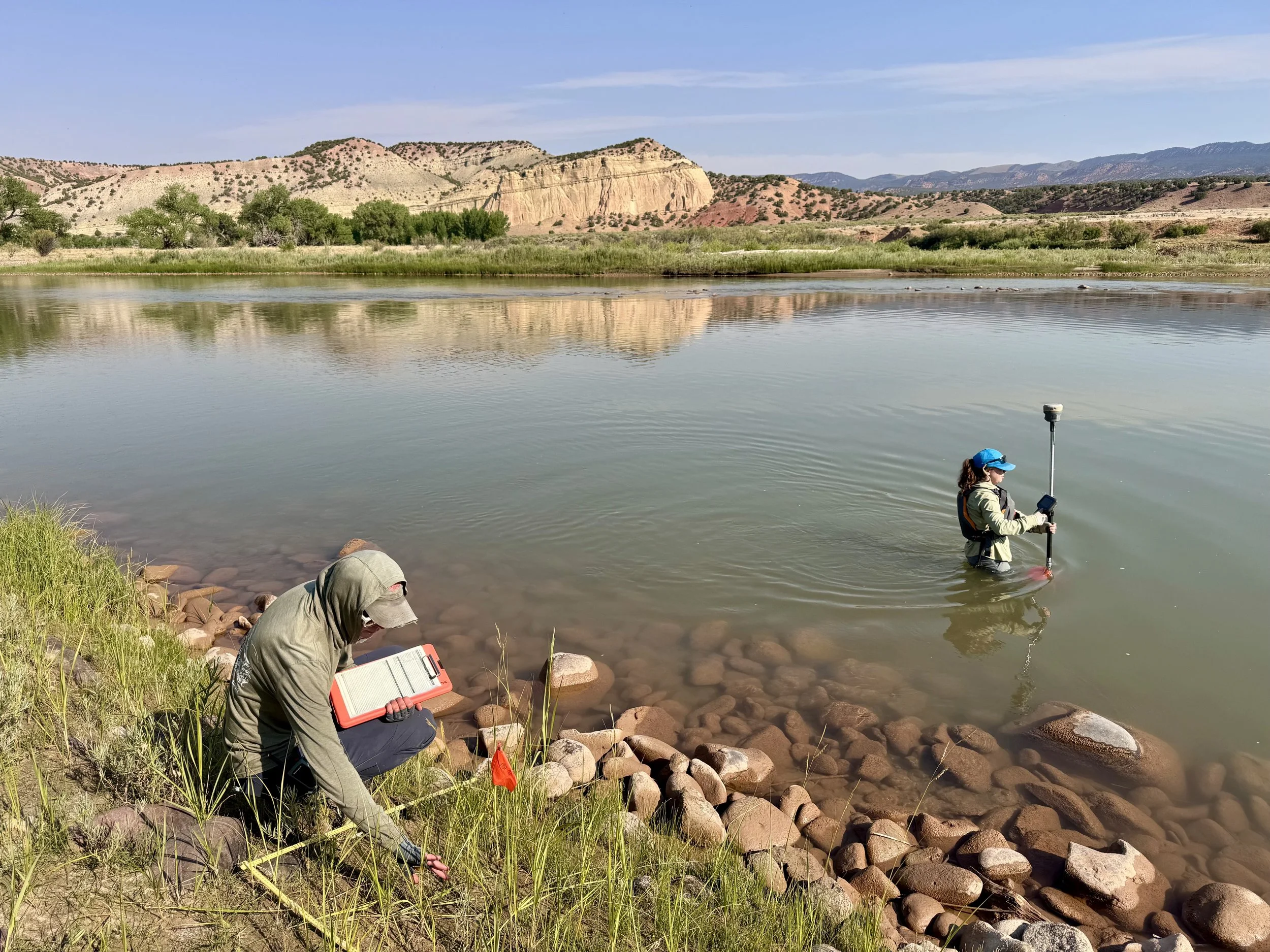 A woman wearing a baseball cap kneels next to a riverbank full of rocks. She is holding a red clipboard.