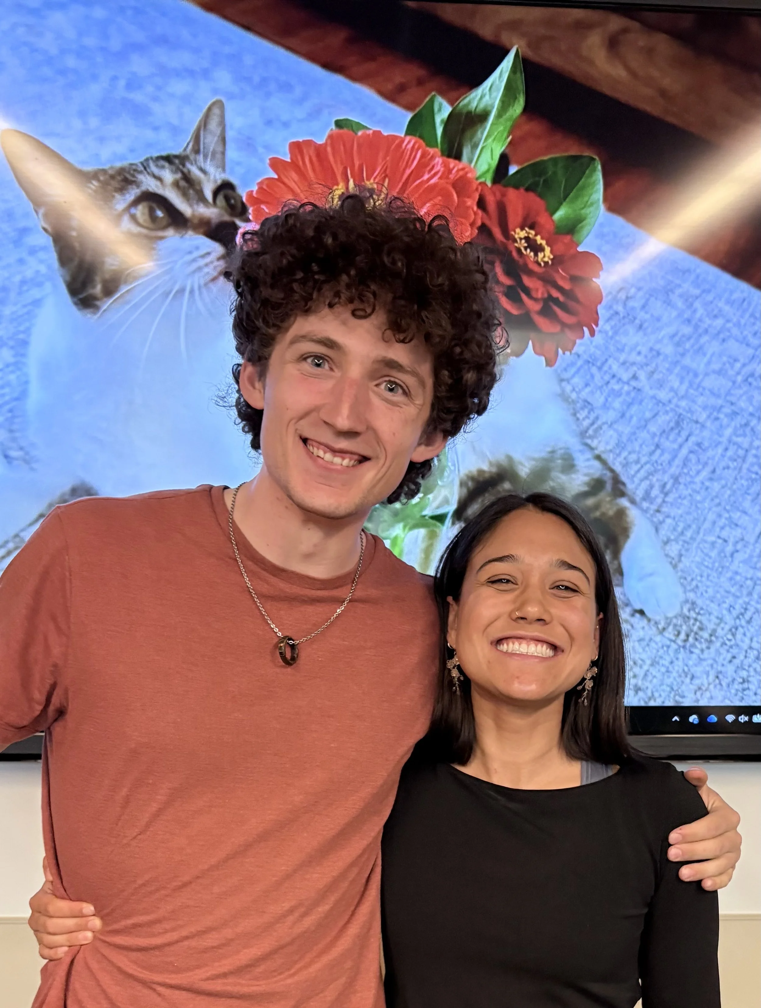 A man with curly hair and a woman with dark brown straight hair stand in front of a TV display monitor with a cat smelling a flower displayed. They are both smiling.