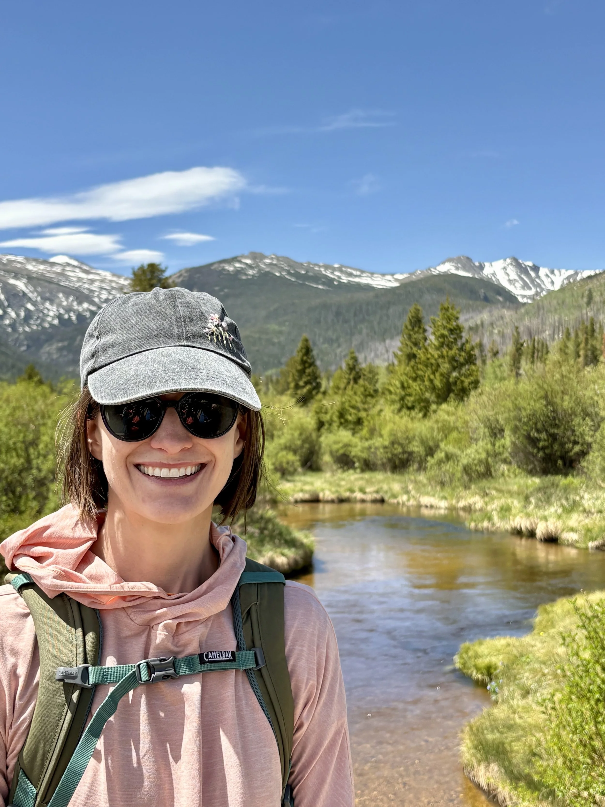 A woman wearing a charcoal baseball cap, sunglasses, and a pink sun shirt stands in front of a mountain creek and snowy mountains