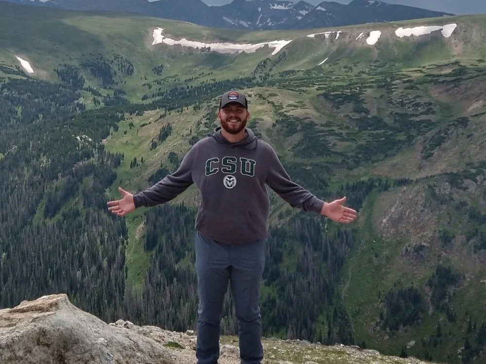 A man wearing a baseball cap and a CSU sweatshirt standing in front of a mountain valley
