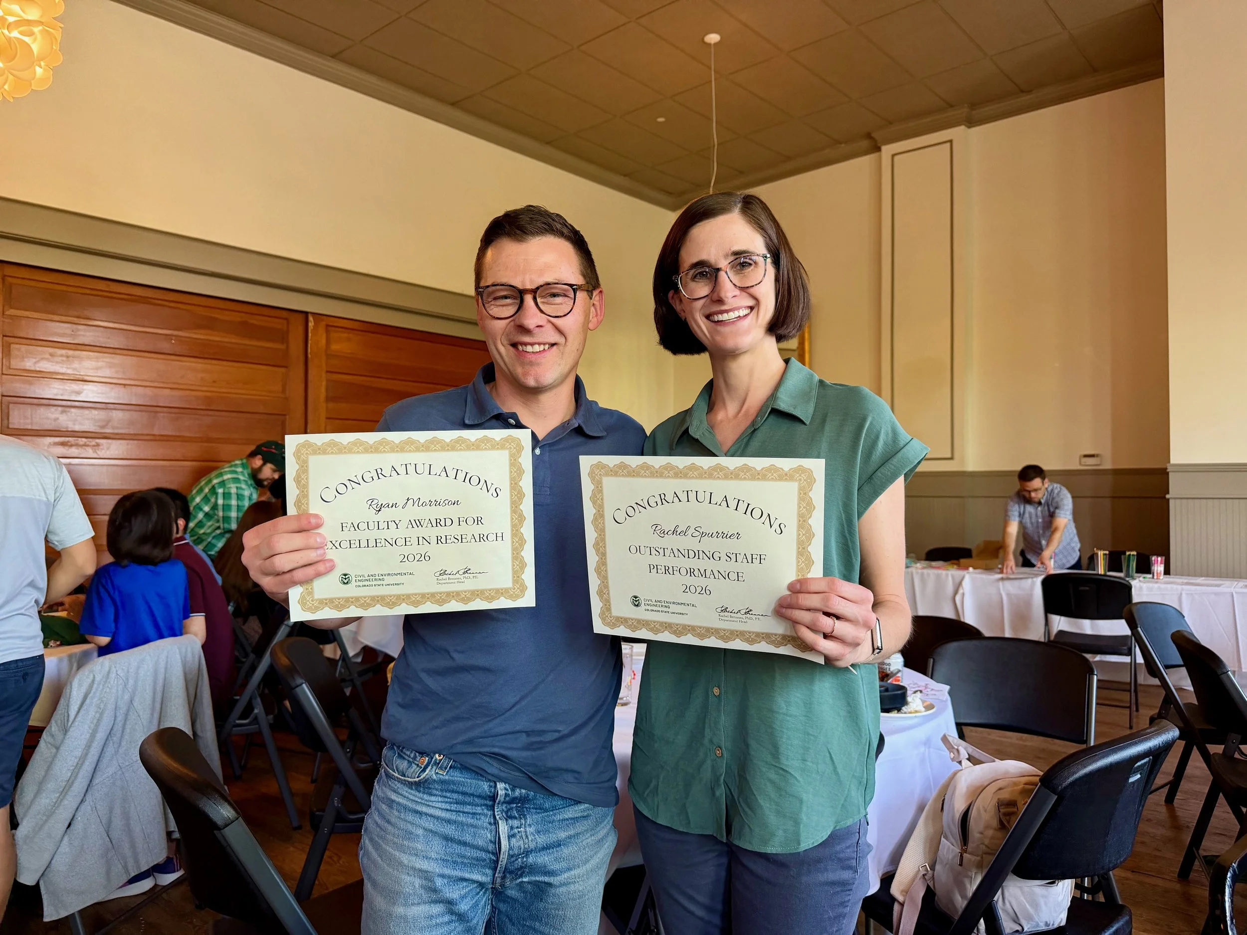A man and a woman both wearing glasses stand next to each other holding paper award certificates.