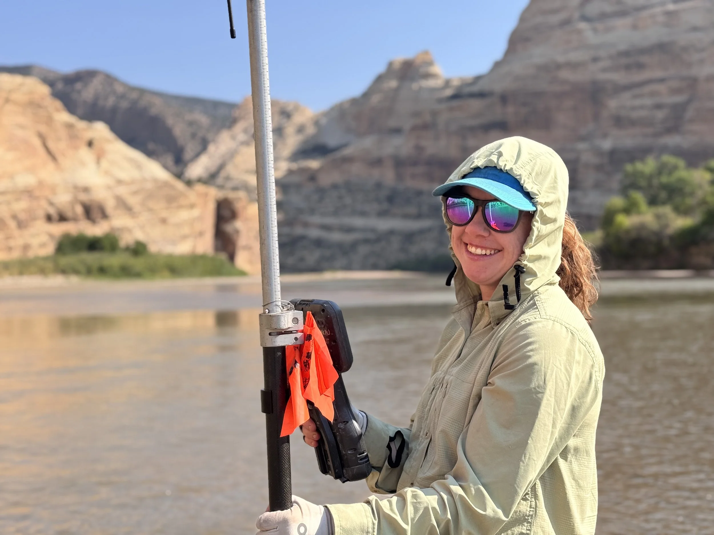 A photograph of a smiling woman with a river and cliff bluffs in the background. She is wearing sunglasses, a baseball cap, and a hooded sun shirt