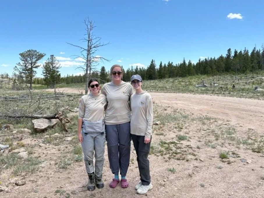 Three women stand in front of a dirt road wearing matching sun shirts