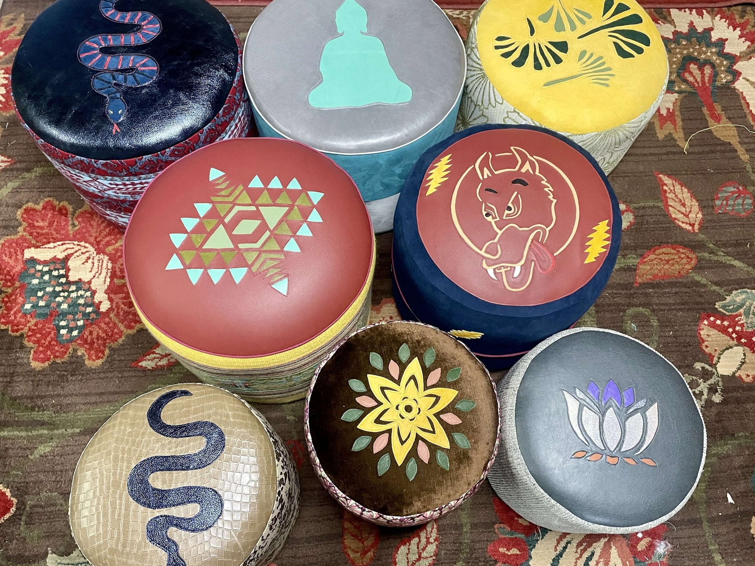 Collection of handcrafted custom ottomans with hand-stenciled designs including snake, Buddha, geometric, and lotus motifs, displayed on a decorative rug