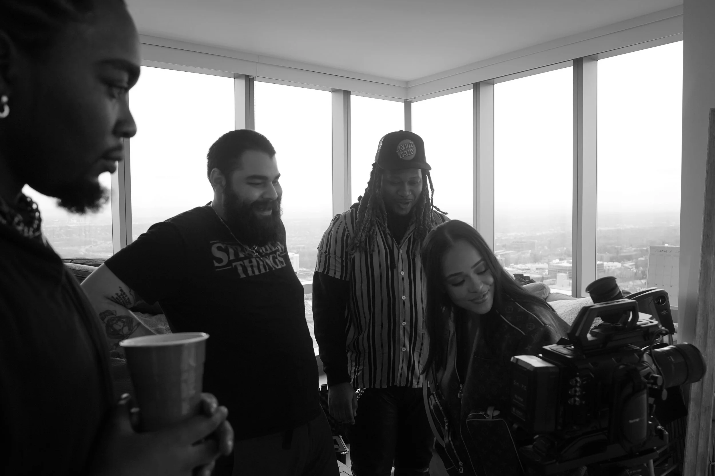 A diverse group of people stand around a camera and smile as they look back at the footage.