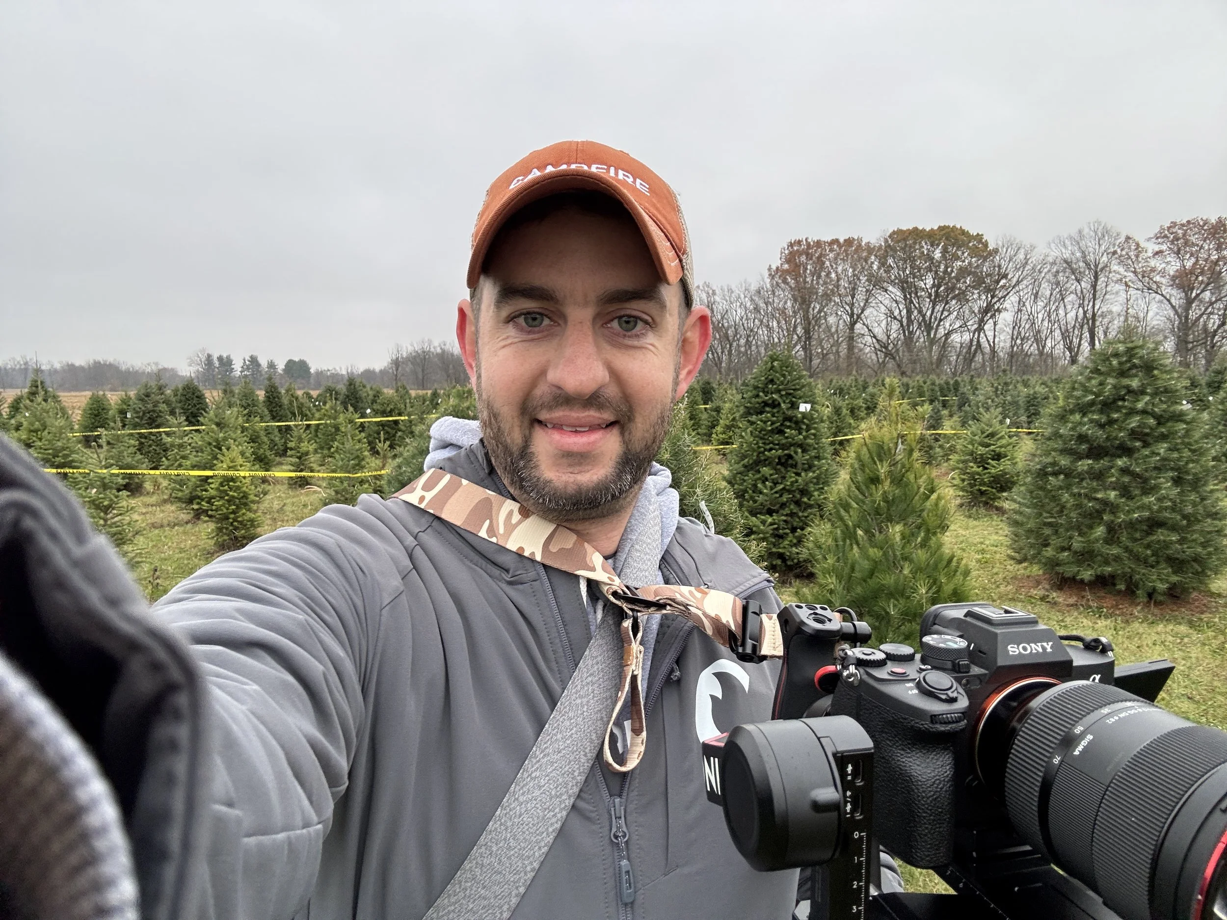 A man taking a selfie at a Christmas tree farm, wearing a gray jacket, orange cap, and carrying a camera.