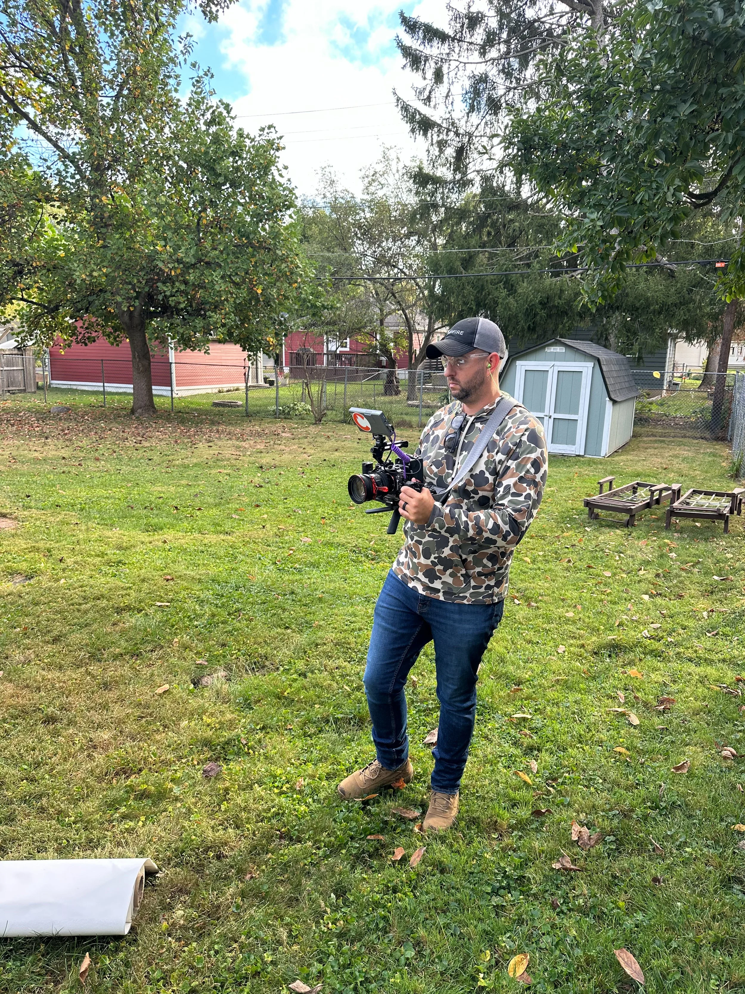 A man in a camouflage hoodie, blue jeans, and tan boots is standing in a backyard, holding a professional camera with a mounted monitor and other equipment. The backyard has green grass, trees, a small shed, and outdoor furniture frames.