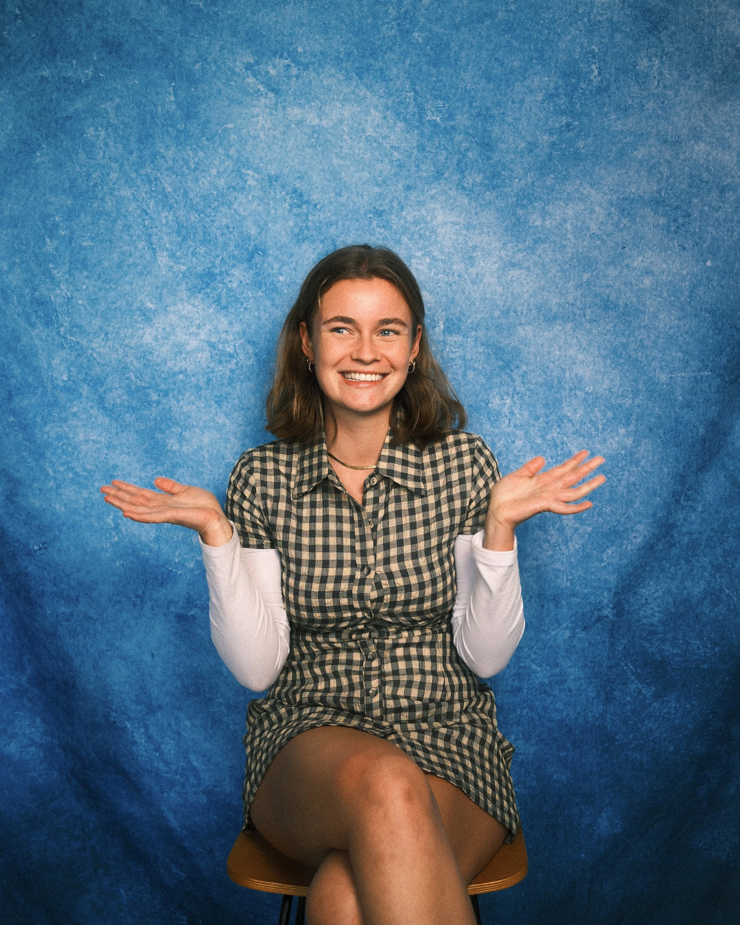 A young woman with long brown hair, wearing a checkered dress and white long sleeve shirt, smiling and sitting on a mustard-colored chair against a blue textured wall. She's raising her hands with palms facing up.