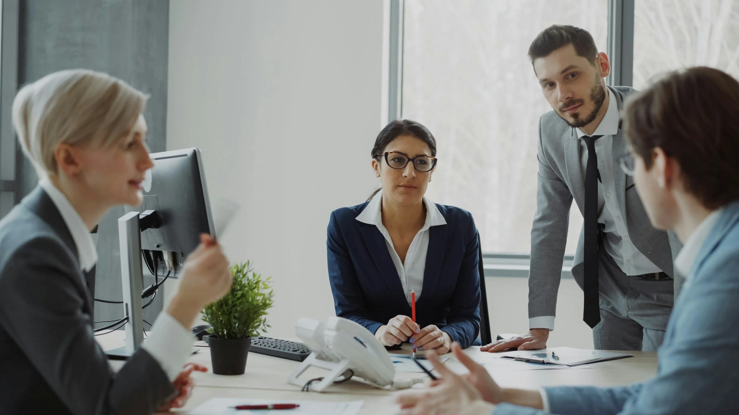 A business meeting with five professionals around a desk in an office with large windows.