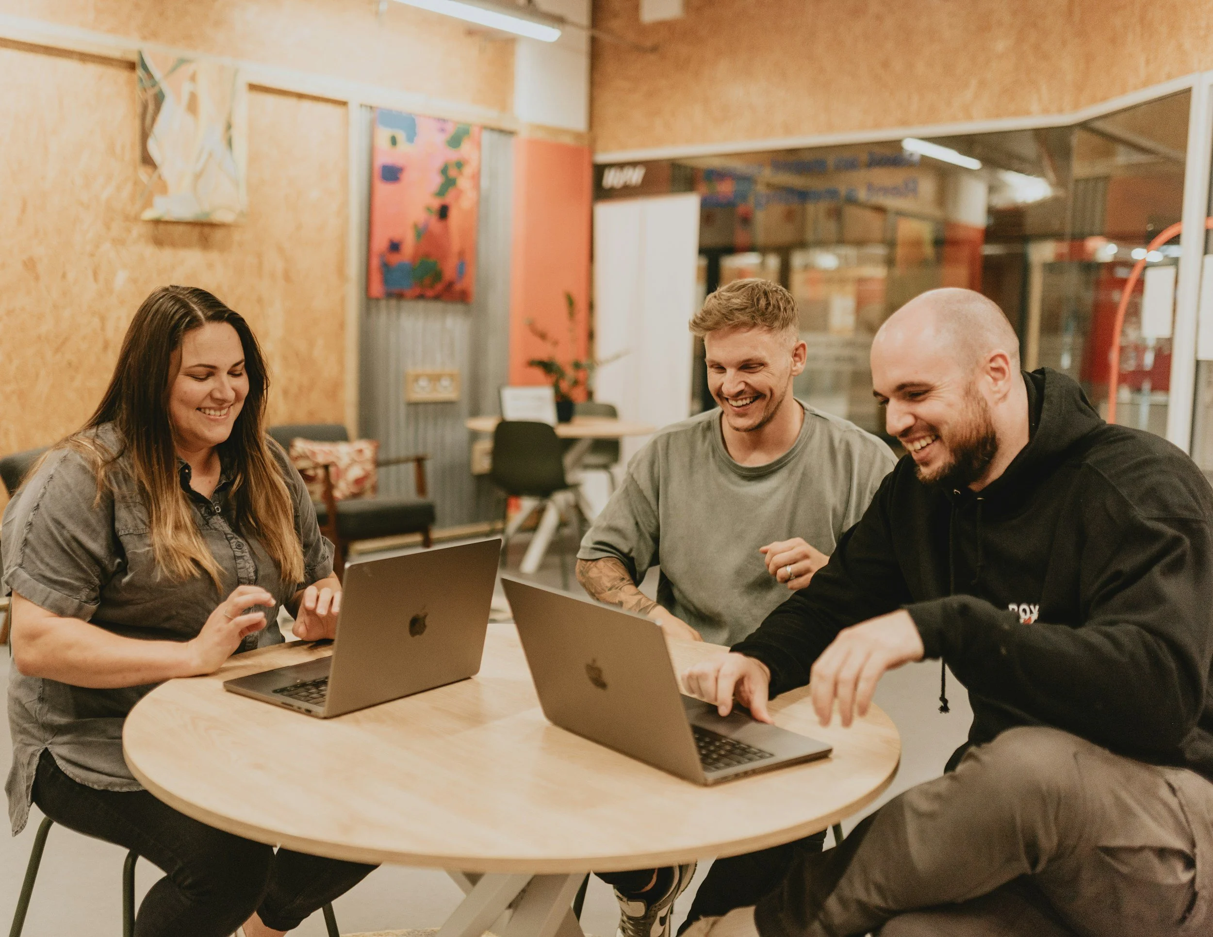 Four friends sitting around a round table with laptops, smiling and enjoying each other's company in a cozy indoor setting with artwork on the wall.