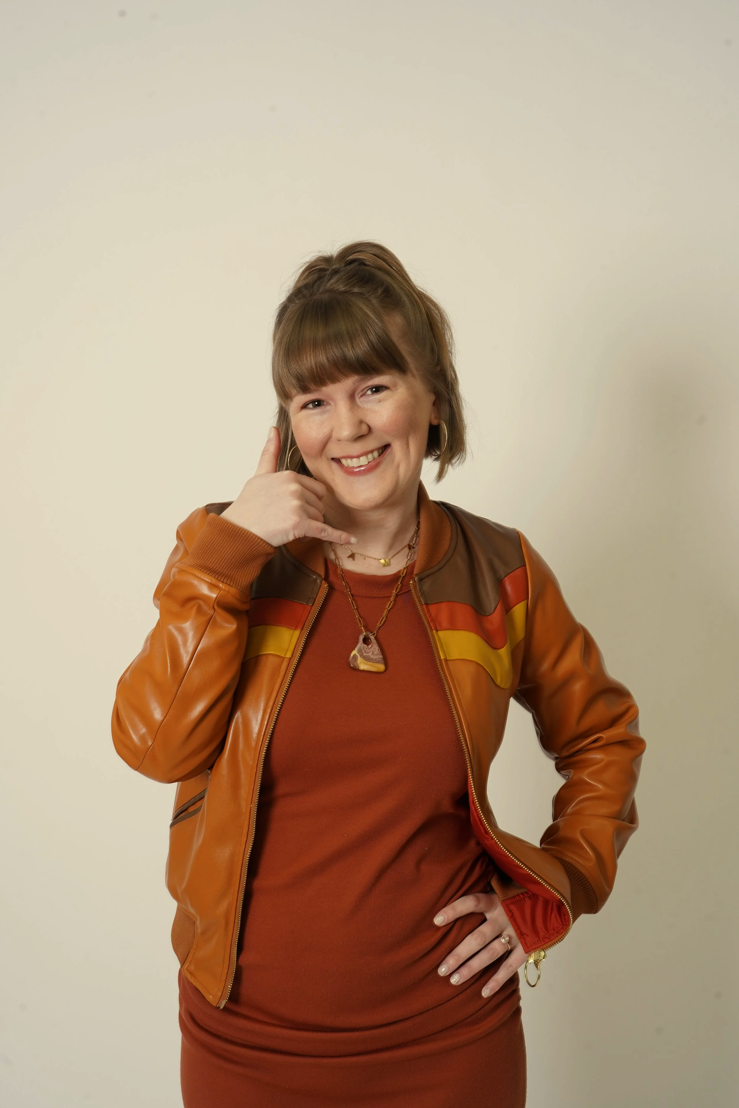 A woman with short brown hair wearing a brown and orange jacket and a matching dress, smiling and gesturing with her right hand while standing against a plain light-colored background.