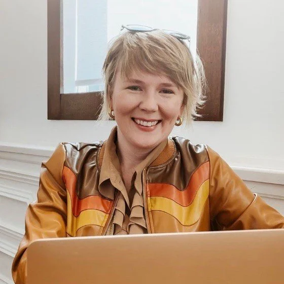 Woman with short blonde hair smiling, wearing a brown and orange jacket, sitting at a table with a laptop in front of her.