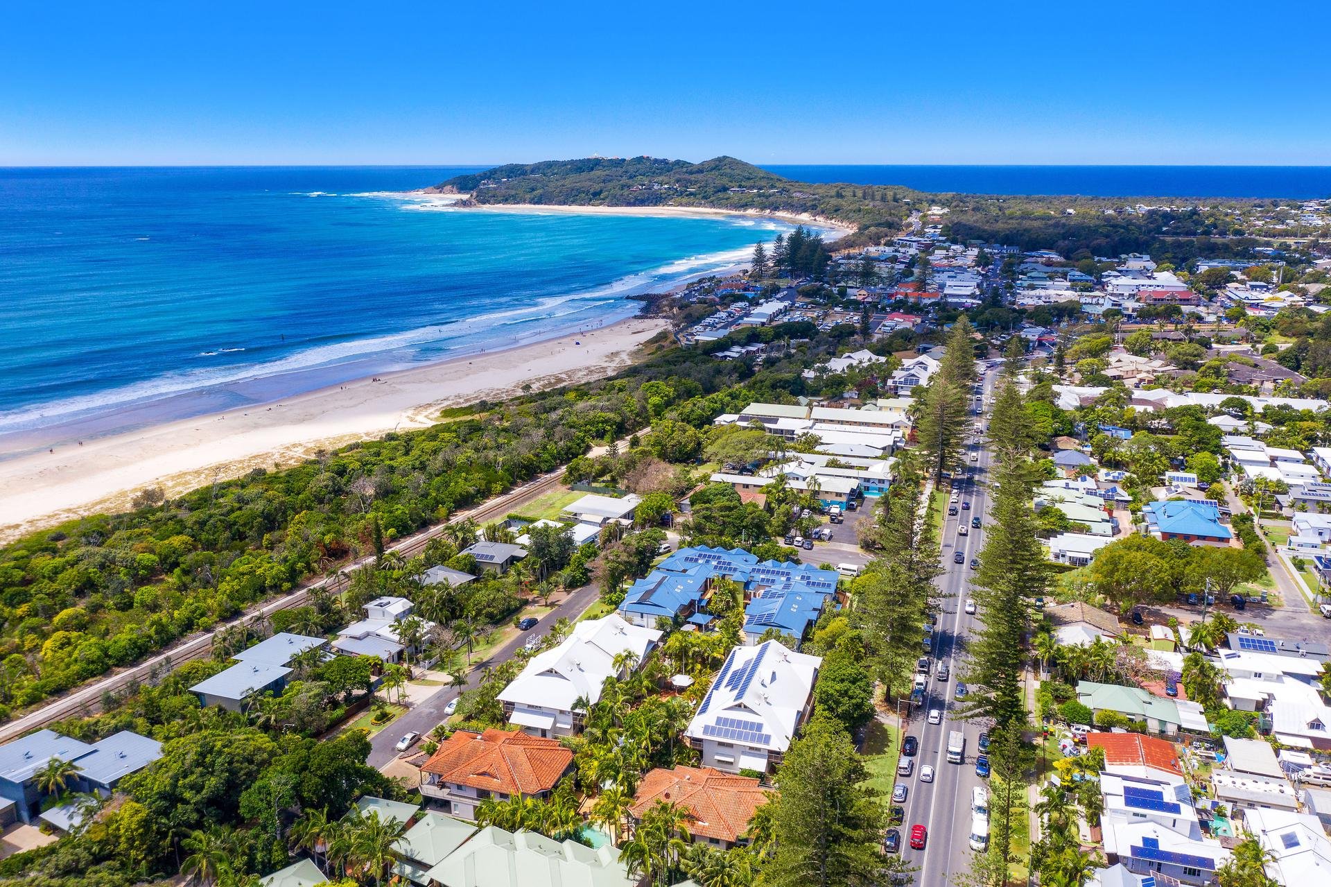 Aerial view of a coastal town with white houses, many with solar panels, lined by a highway with cars, green trees, and a sandy beach leading to the ocean with clear blue water, in a sunny climate.