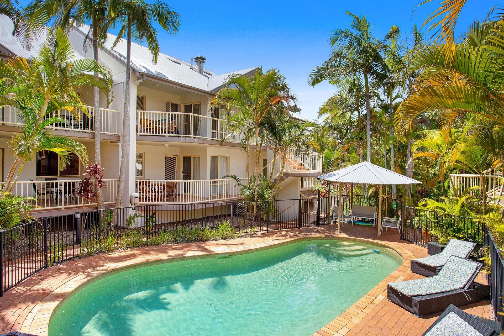 A tropical apartment complex landscaped with palm trees, featuring a swimming pool with lounge chairs, and a multi-story building with balconies.