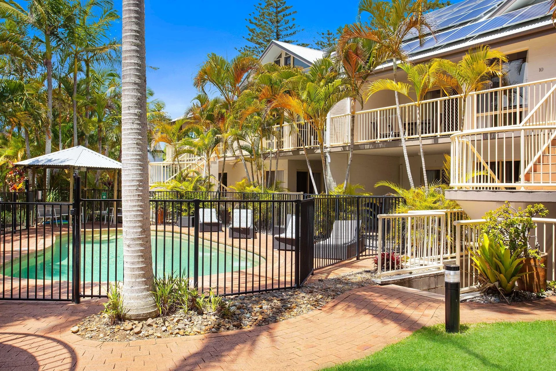 Residential apartment complex with balconies, surrounded by tropical palm trees, with a swimming pool area enclosed by a black metal fence and lounge chairs. Solar panels are visible on the roof under a clear blue sky.