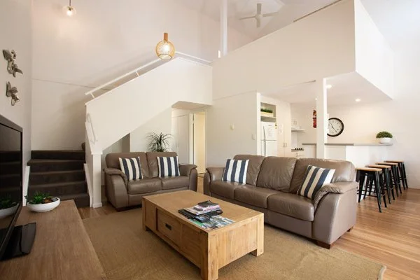 Living room with brown leather sofa, matching armchair, wooden coffee table, staircase leading upstairs, TV on stand, and an open kitchen with bar stools in the background.