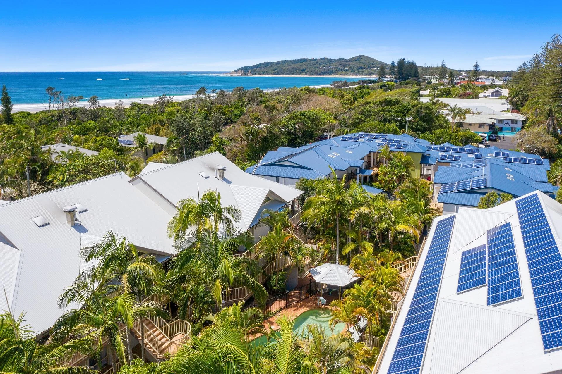 Aerial view of houses with solar panel roofs surrounded by lush green trees, overlooking a beach with blue ocean and a distant hill under a clear blue sky.