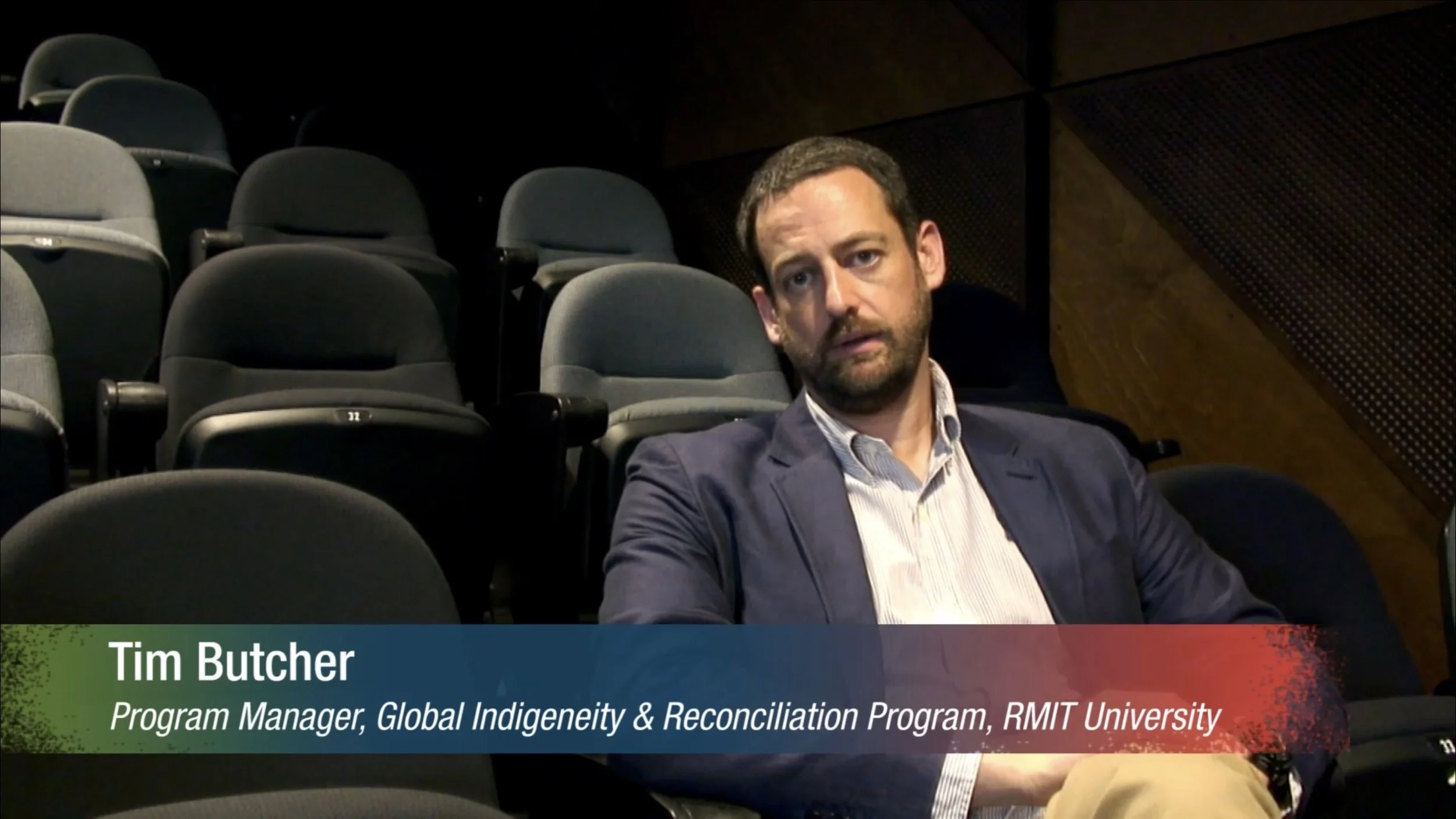 A man in a navy blazer and striped shirt sitting in an empty auditorium or lecture hall with black and gray seats. There is a lower third graphic displaying his name, Tim Butcher, and his title, Program Manager, Global Indigeneity & Reconciliation Program, RMIT University.