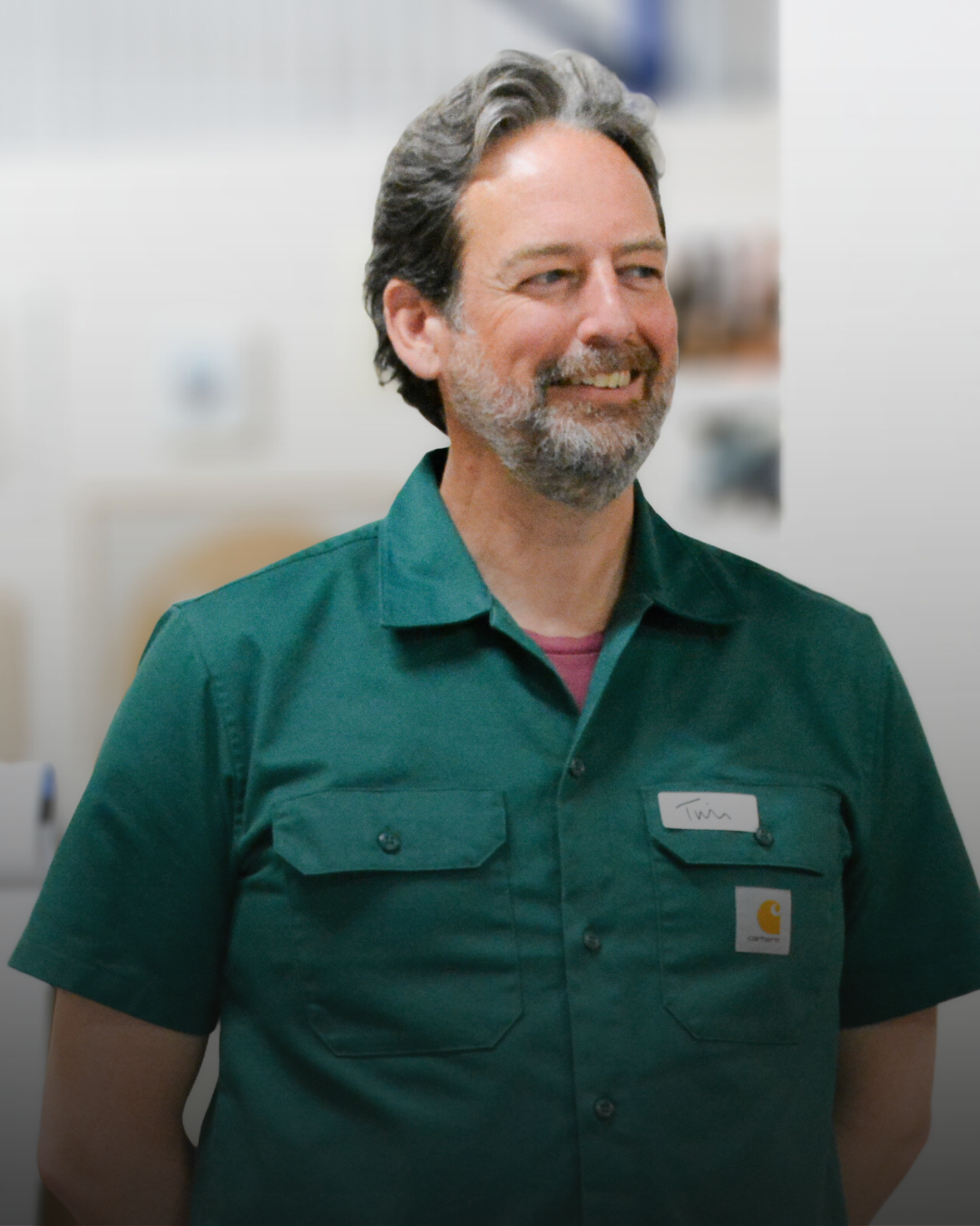 A smiling man with gray hair and beard, wearing a green work shirt with a Carhartt logo and a name tag that reads 'Tina', standing in an indoor work environment.