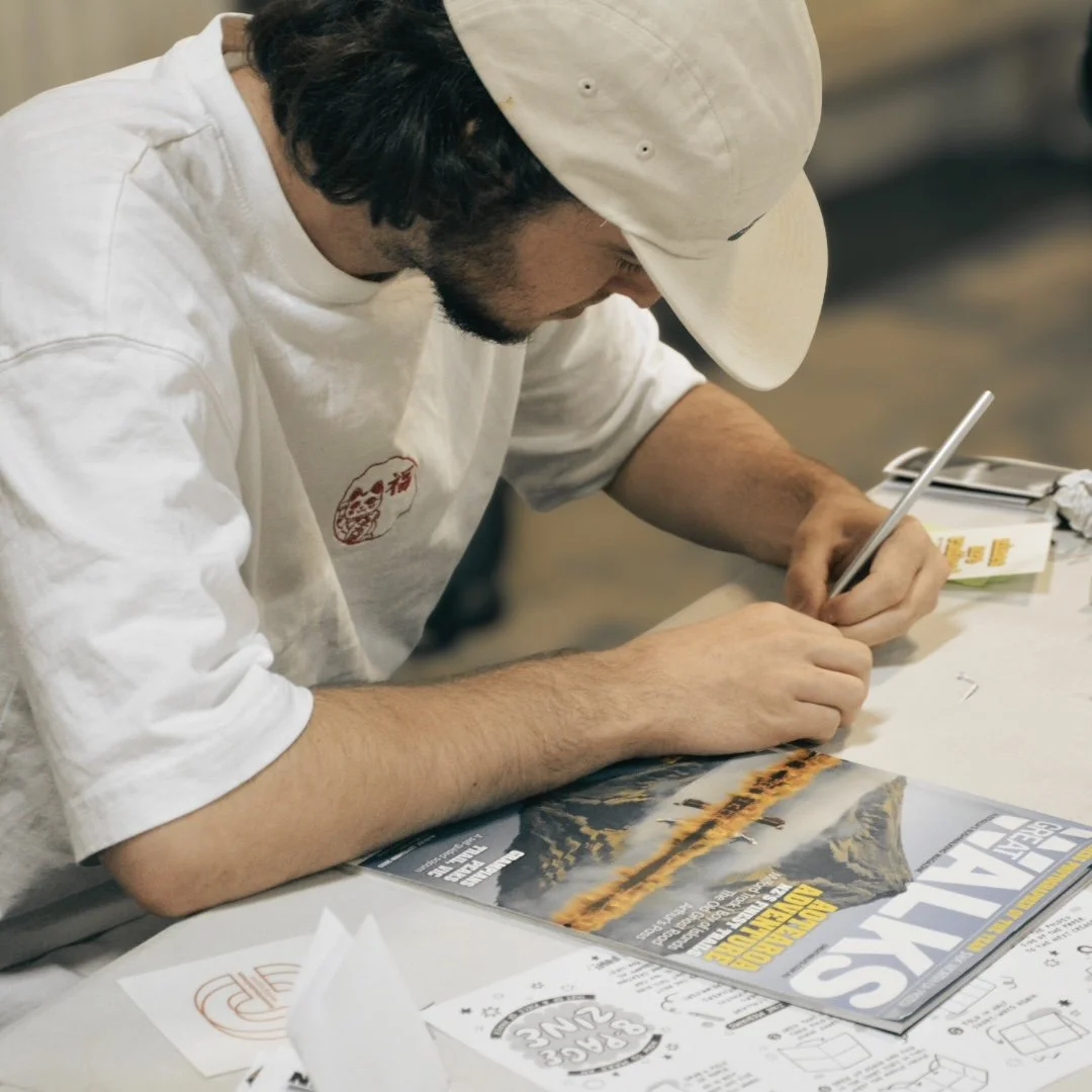 A man wearing a white cap and shirt is sitting at a table, signing a poster with a marker. There are papers and a magazine on the table.