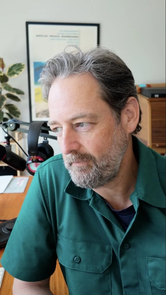 A man with gray hair and a beard wearing a green shirt, sitting at a desk with a computer and headphones, in a home office with framed artwork and a plant in the background.