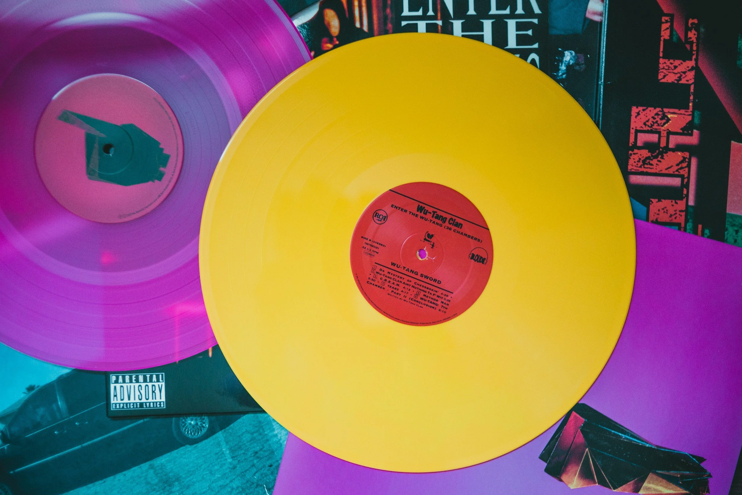 Colorful vinyl records, including a yellow one with a red label featuring Wu-Tang Clan and Wu-Tang Sword, are displayed on a surface along with partially visible album covers in the background.