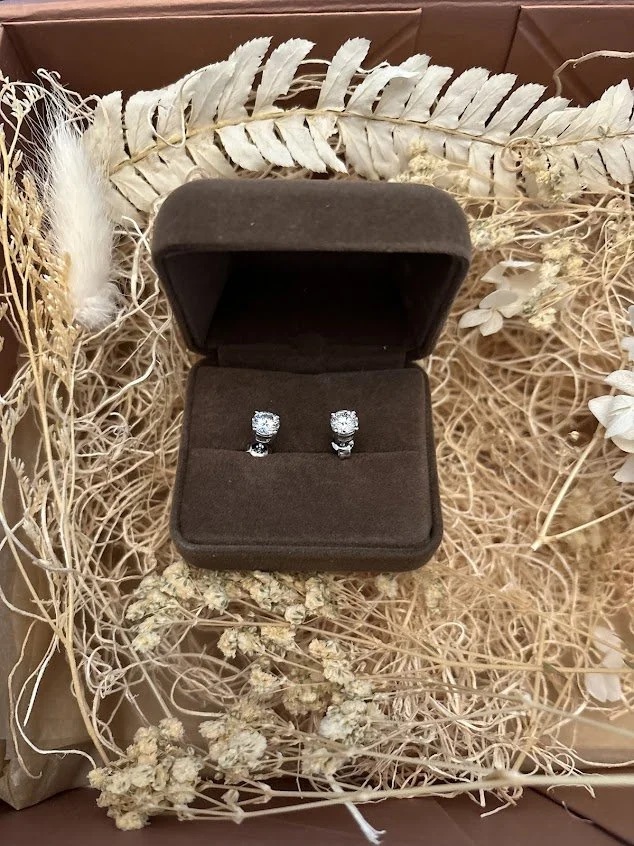 A pair of sparkling diamond earrings resting in a soft velvet box, nestled in a bed of dried floral elements.