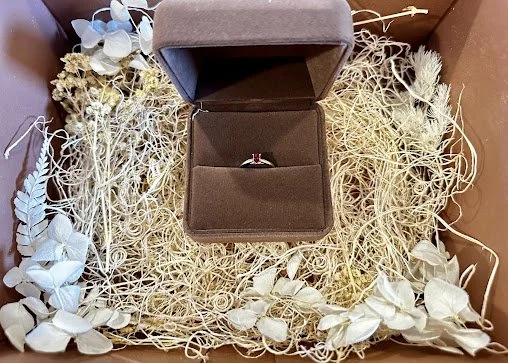 A beautifully boxed memorial ring with a delicate red stone, nestled in an elegant display case surrounded by dried flowers.