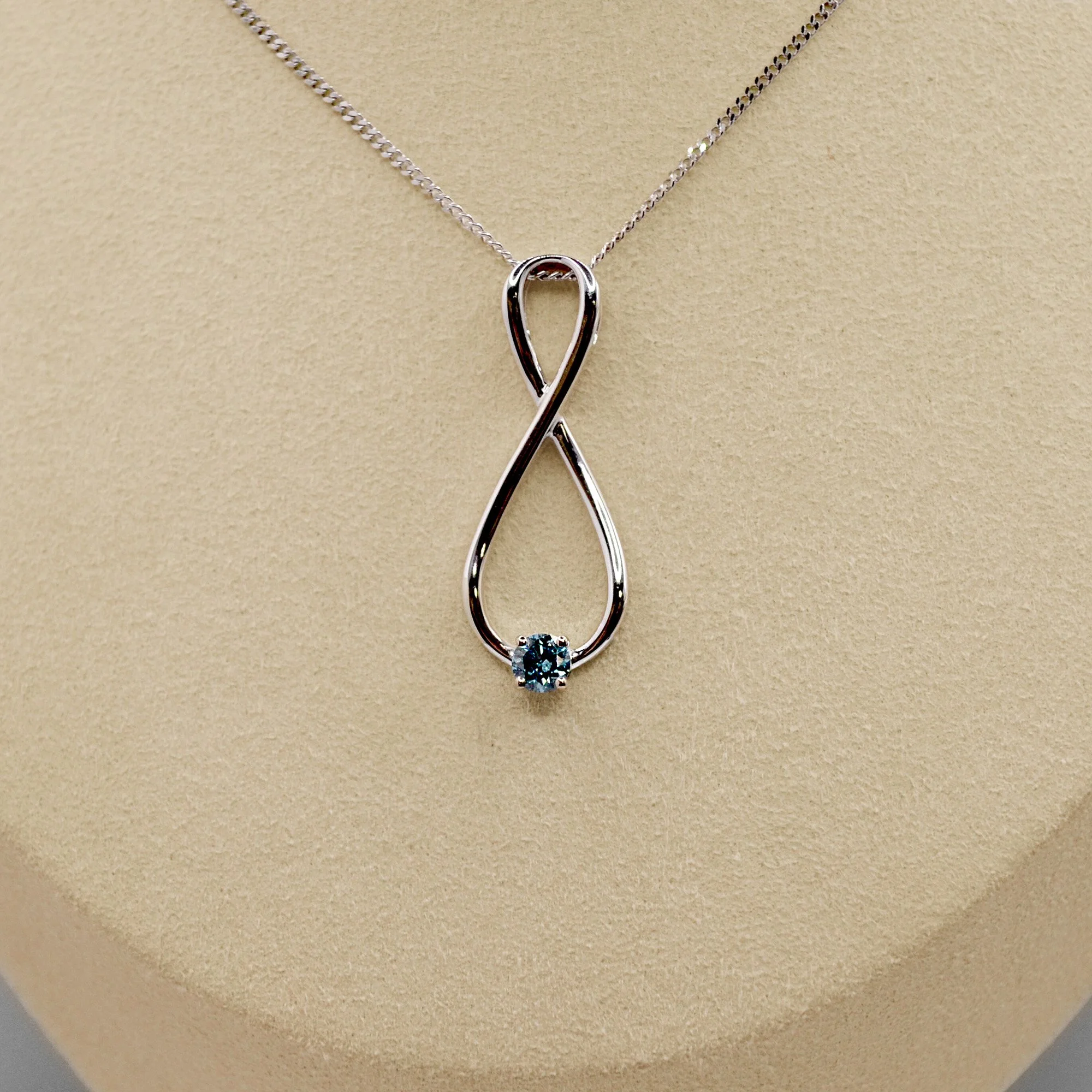 Infinity-shaped platinum pendant adorned with a small blue diamond at the bottom, suspended on a fine platinum chain.