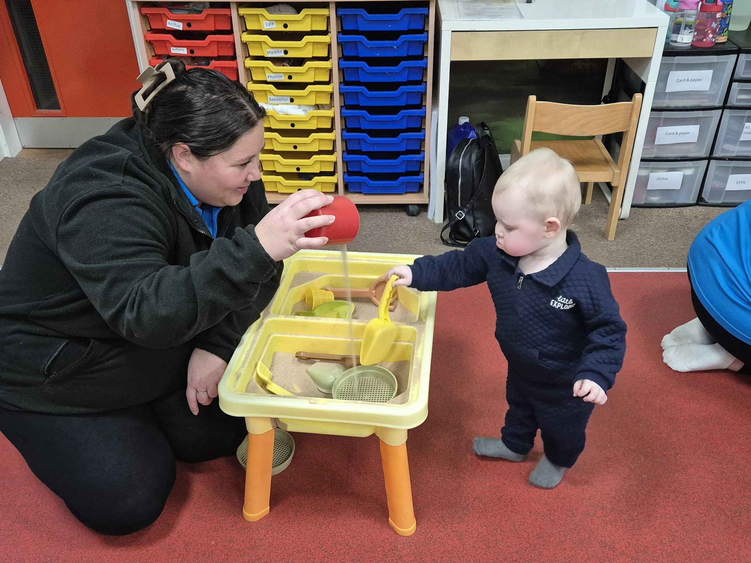 A woman and a young boy playing at a water table in a classroom. The woman is pouring water into the table while the boy is holding a yellow toy with his right hand. The background shows colorful storage bins and classroom furniture.