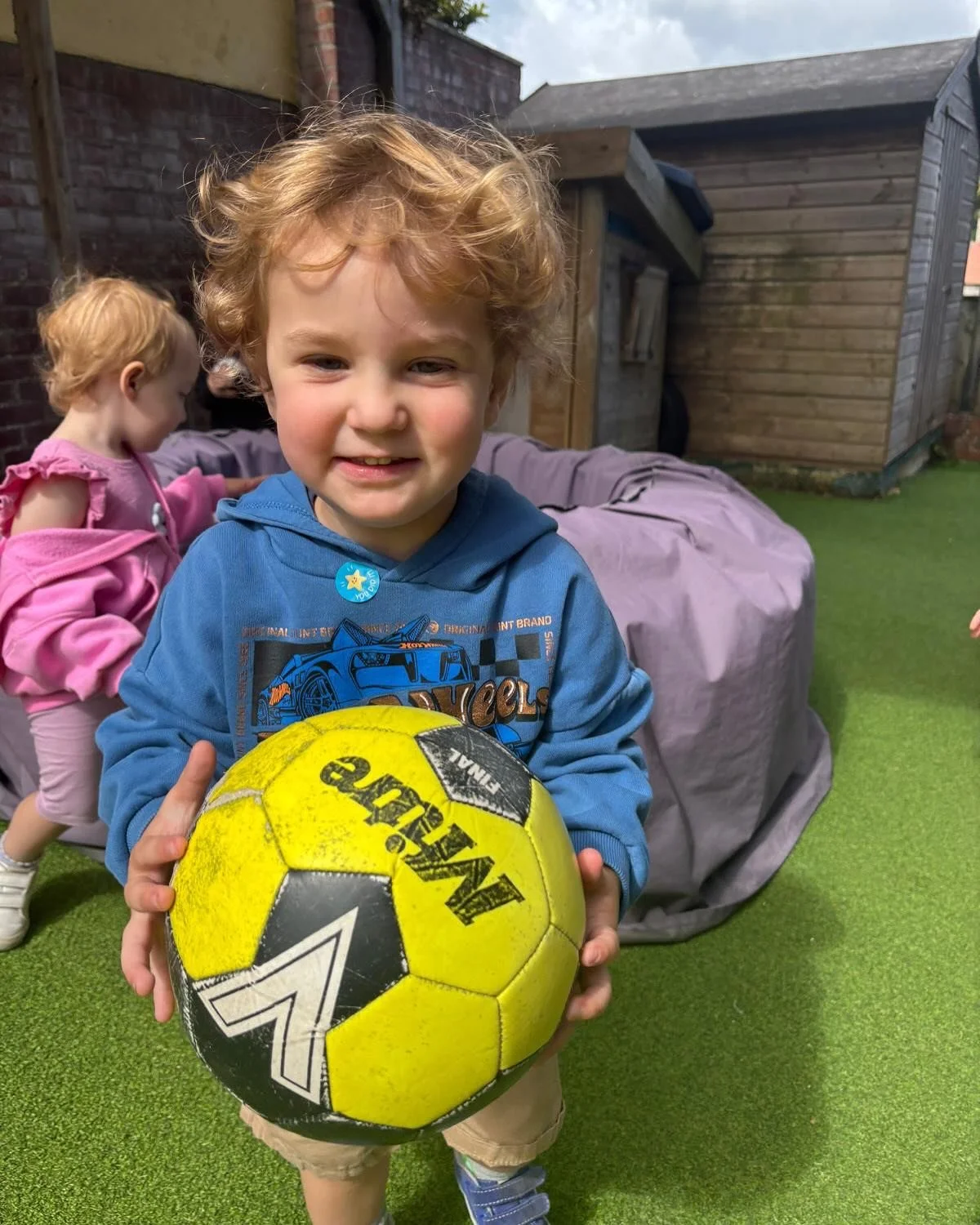 A young boy with curly red hair smiling and holding a yellow and black soccer ball in an outdoor backyard. In the background, a young girl wearing pink and a purple cover is playing near a covered object and a wooden shed.