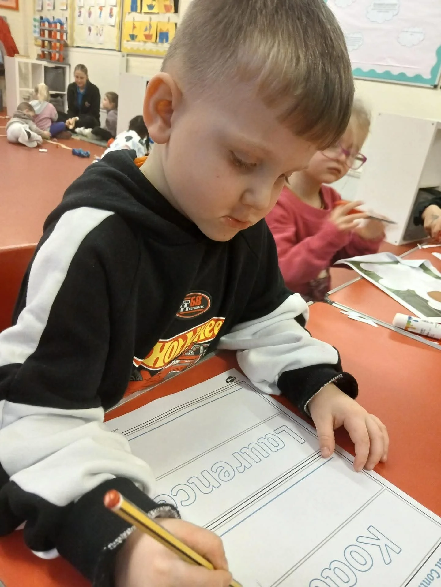 Child writing in a workbook in a classroom with other children and a teacher in the background.