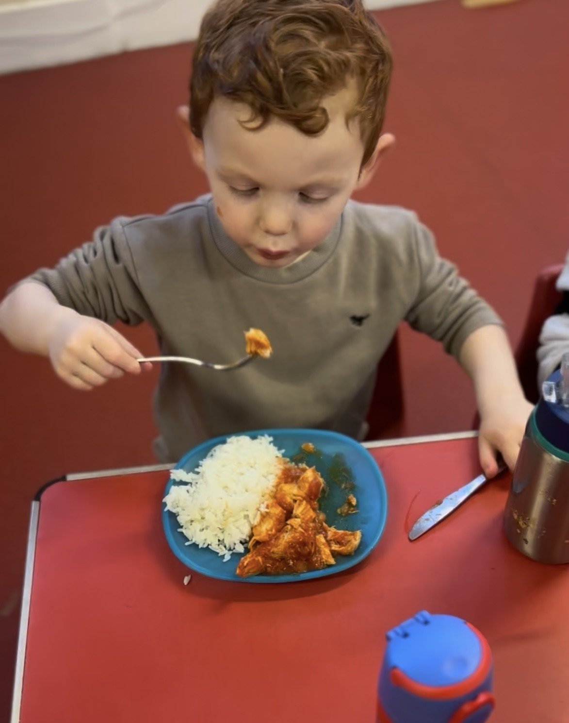 A young boy with curly red hair eating food from a blue plate, which contains rice and a stew or curry, at a red table.
