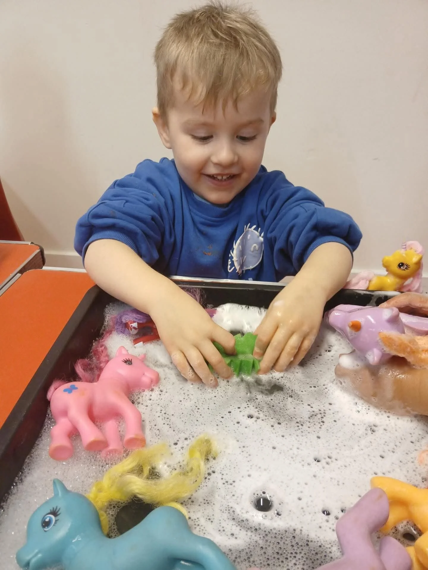 A young boy with blond hair washing plastic toy ponies in a soapy water bath at a play area.