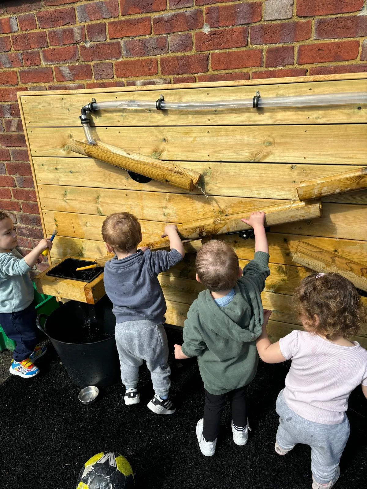 Four children are playing with water on a wooden water wall outside near a brick wall. The children are standing and using their hands to interact with the water flowing down the wall. There is a soccer ball on the ground near them.