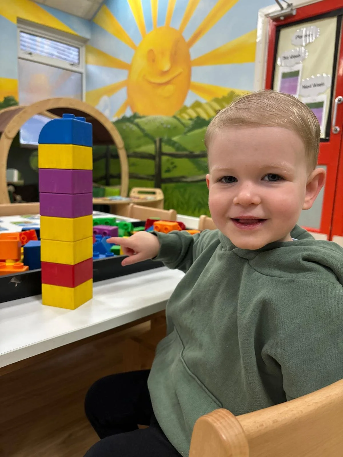 A smiling young boy with blonde hair sits at a table in a colorful playroom, pointing at a tower of large colorful building blocks, with a mural of a smiling sun and green hills in the background.