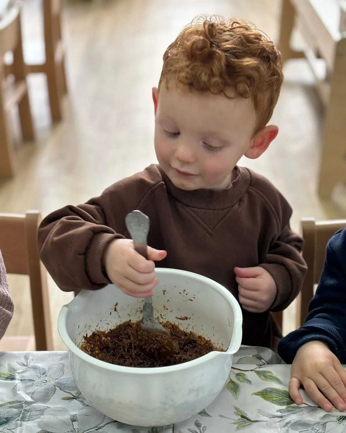 A young boy with curly red hair stirring a mixture in a white bowl with a spoon, sitting at a table with a floral tablecloth, in a room with wooden chairs.
