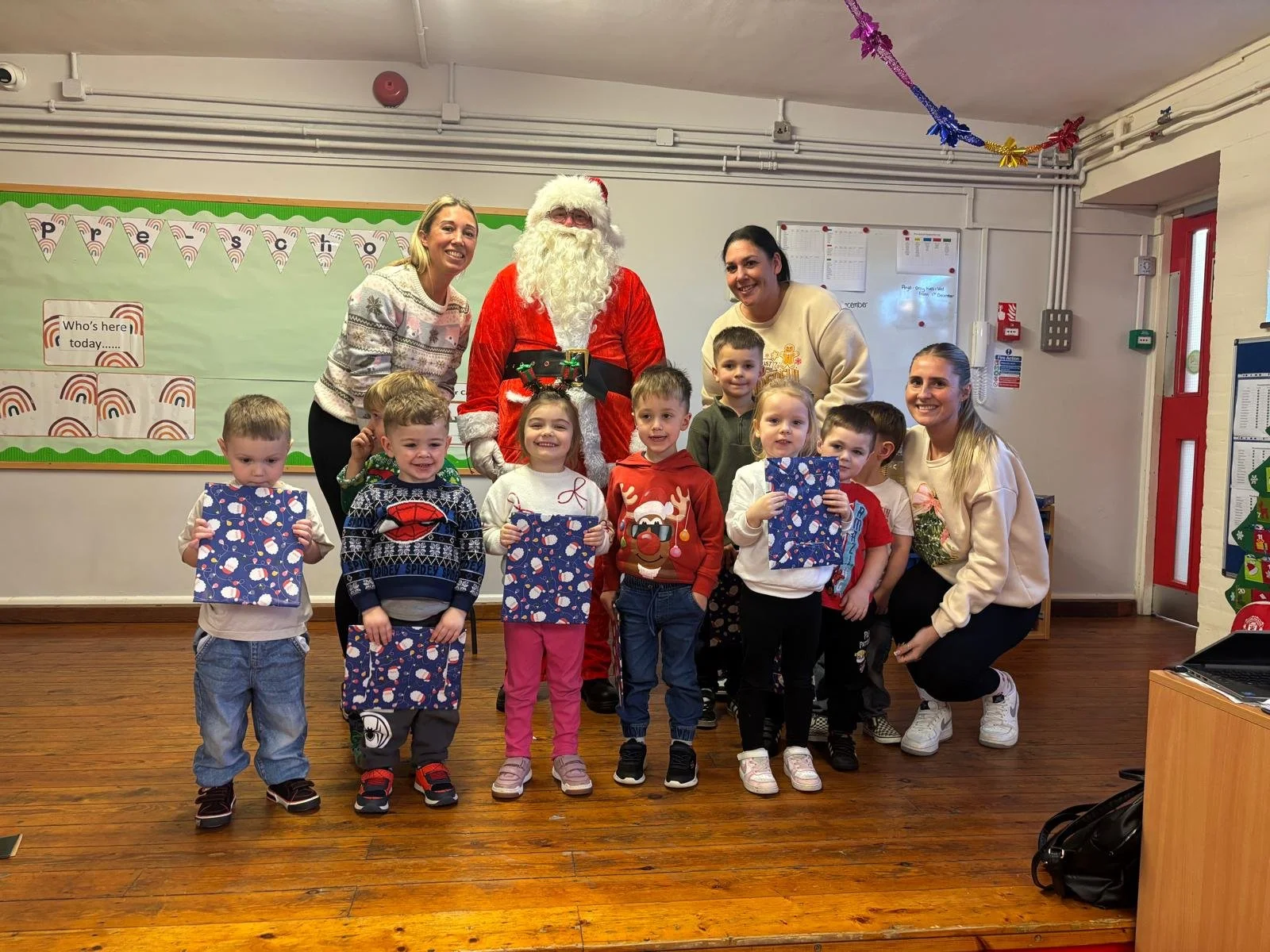 Children holding wrapped Christmas presents with Santa Claus and three women in a decorated classroom.
